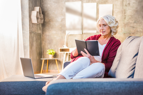 Elderly woman reading book comfortably on sofa with laptop nearby, enjoying clear vision post-cataract treatment