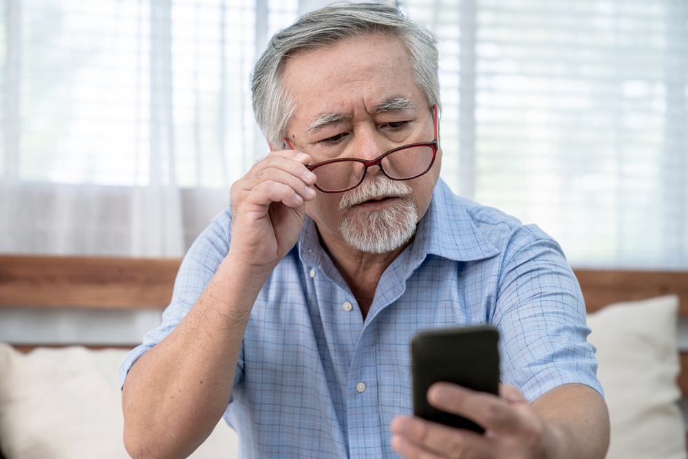 Asian elderly man adjusting glasses to read phone on bed, struggling with blurry vision from presbyopia or early cataracts